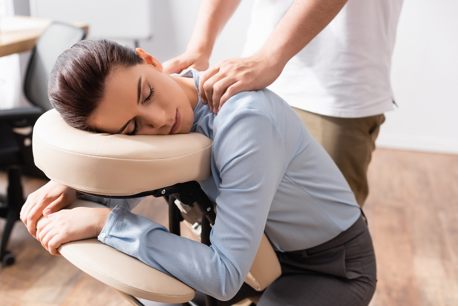 Massage therapist massaging shoulders of woman with closed eyes, sitting on massage chair with office on blurred background